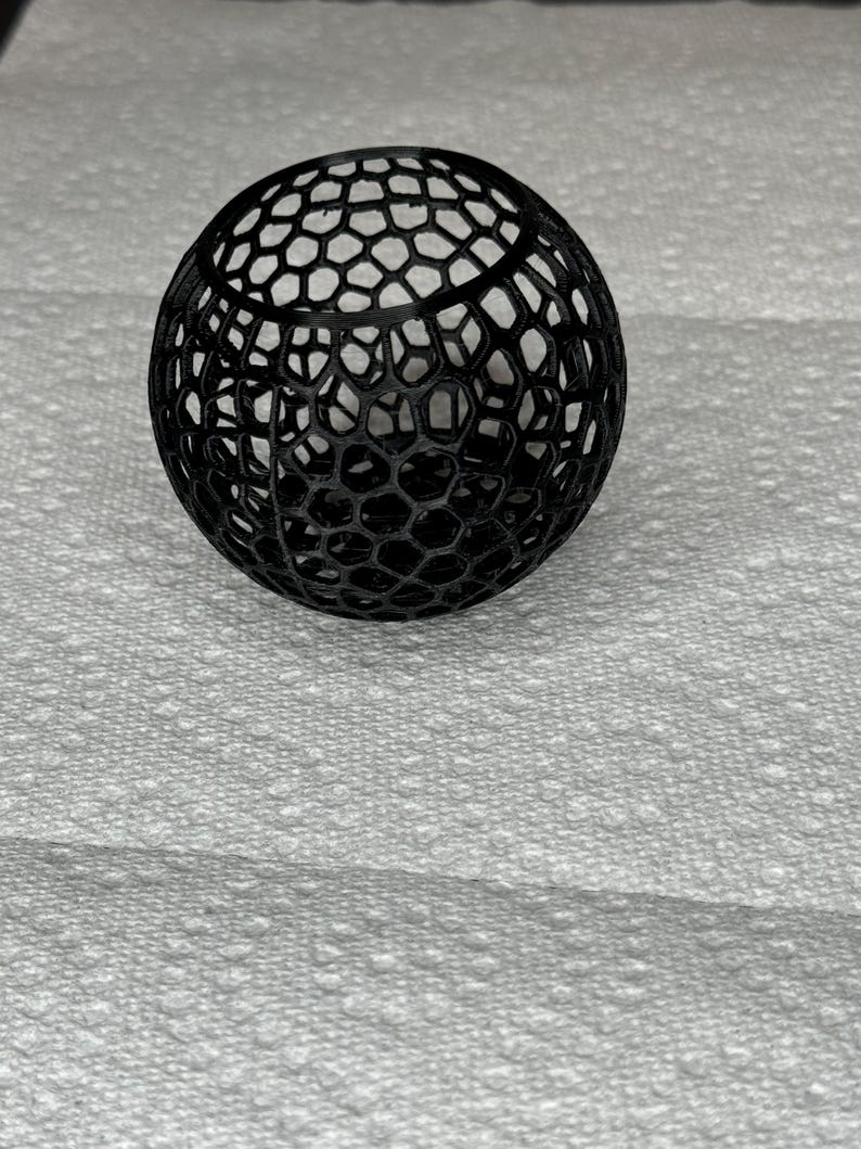 May include: A black, spherical object with a honeycomb-like pattern. The intricate design features a series of interconnected hexagonal shapes, creating an open, lattice-like structure. The object is likely a decorative item or a component of a larger design.