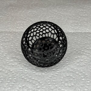May include: A black, spherical decorative object with a honeycomb-like pattern. The object is made of a series of interconnected shapes, creating an open, airy design. The sphere sits on a white, textured surface.