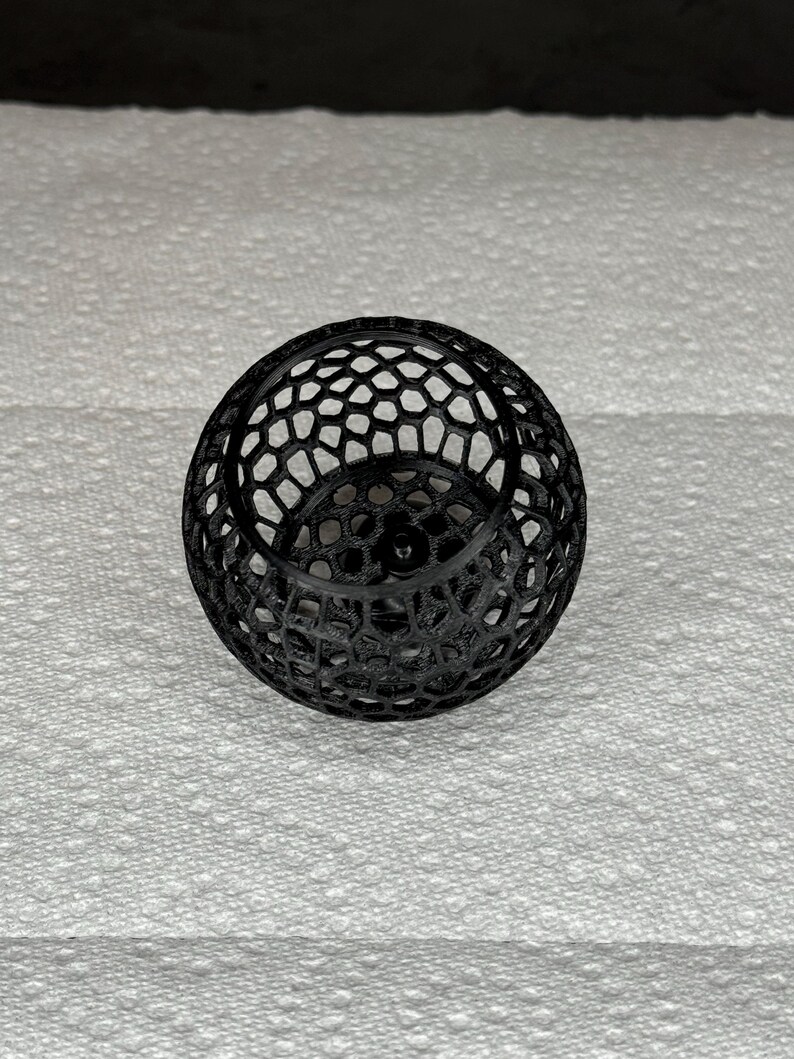 May include: A black, spherical decorative object with a honeycomb-like pattern. The object is made of a series of interconnected shapes, creating an open, airy design. The sphere sits on a white, textured surface.