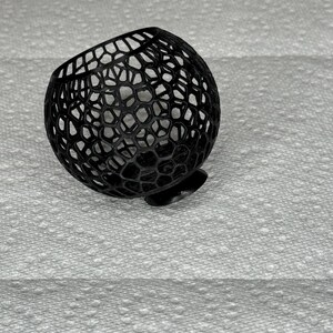 May include: A black, spherical decorative object with a honeycomb-like pattern. The object is made of a series of interconnected shapes, creating an open, airy design. It has a small, flat base, and is resting on a textured white surface.