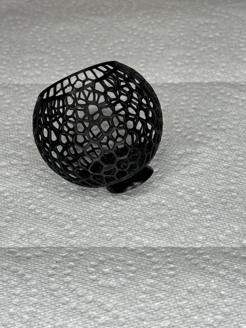 May include: A black, spherical decorative object with a honeycomb-like pattern. The object is made of a series of interconnected shapes, creating an open, airy design. It has a small, flat base, and is resting on a textured white surface.