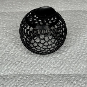 May include: A black, spherical decorative object with a honeycomb-like pattern. The top features a solid, rounded cap. The object is likely 3D-printed and sits on a textured white surface.