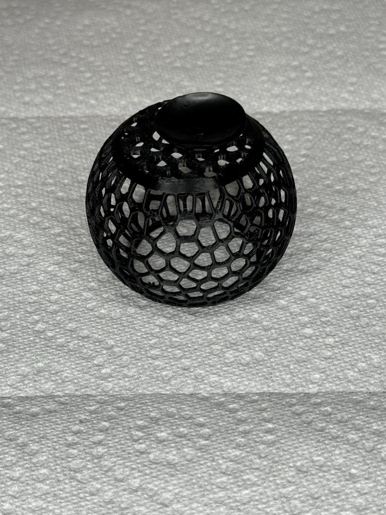 May include: A black, spherical decorative object with a honeycomb-like pattern. The top features a solid, rounded cap. The object is likely 3D-printed and sits on a textured white surface.