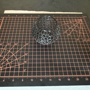 May include: A black, spherical, openwork decorative object. The object has a honeycomb-like pattern and sits on a black cutting mat with orange grid lines and measurements in inches.