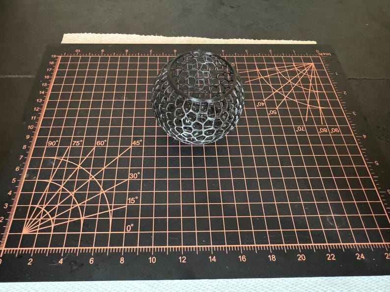 May include: A black, spherical, openwork decorative object. The object has a honeycomb-like pattern and sits on a black cutting mat with orange grid lines and measurements in inches.