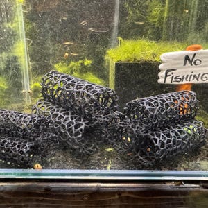 Può includere: Primo piano di una decorazione per acquario in rete nera. La decorazione ha la forma di un cilindro ed è impilata su se stessa. La decorazione è in un acquario con piante verdi e un cartello che dice "No Fishing!!".
