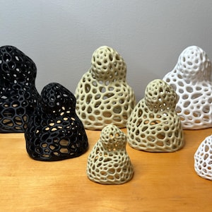 May include: A collection of decorative ceramic sculptures in black, off-white, and beige. The sculptures have a unique, open-weave design, resembling organic forms. The pieces vary in size and are arranged on a wooden surface.