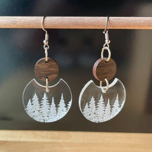 May include: A pair of dangle earrings. Each earring features a dark brown wooden circle above a clear, crescent-shaped acrylic piece. The acrylic has a white etched forest design. The earrings hang from silver-colored hooks.