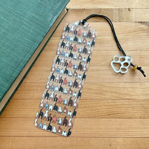 May include: A clear bookmark featuring a repeating pattern of dog rears in various colors. The bookmark has a black cord and a silver paw print charm with a small yellow bead. The bookmark is next to a green book on a wooden surface.