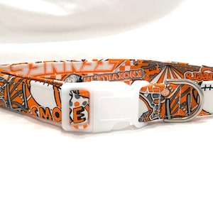 Puede incluir: Collar de perro naranja y blanco con una hebilla de pl&aacute;stico blanca y un anillo en D de metal plateado. El collar presenta un patr&oacute;n de varias im&aacute;genes, incluyendo una pelota de b&eacute;isbol, una pelota de baloncesto, una pelota de f&uacute;tbol y las palabras "Knoxville" y "Smokies".