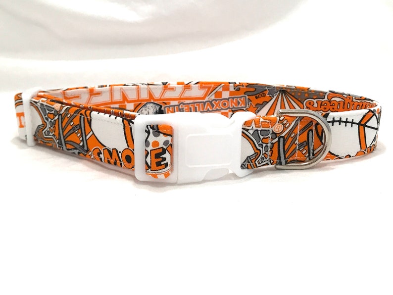 Puede incluir: Collar de perro naranja y blanco con una hebilla de pl&aacute;stico blanca y un anillo en D de metal plateado. El collar presenta un patr&oacute;n de varias im&aacute;genes, incluyendo una pelota de b&eacute;isbol, una pelota de baloncesto, una pelota de f&uacute;tbol y las palabras "Knoxville" y "Smokies".