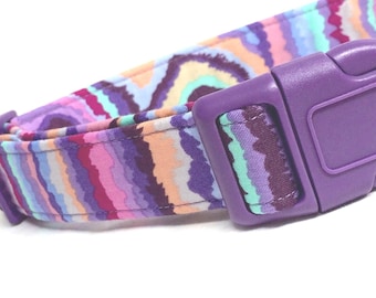 May include: A pet collar featuring a wavy, multi-colored pattern of purple, magenta, light blue, and peach stripes. The collar is equipped with a solid purple plastic side-release buckle and a matching purple plastic adjustment slider.