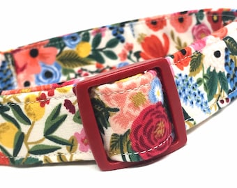 May include: A white dog collar with a floral print and a red buckle. The floral print features red, pink, yellow, blue, and green flowers.