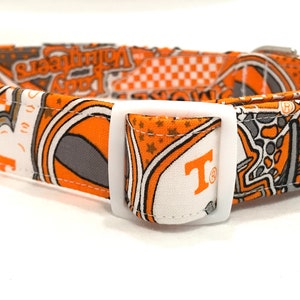 Puede incluir: Collar de perro naranja y gris con una hebilla blanca. El collar tiene un patr&oacute;n de estrellas, tableros de ajedrez y la letra "T" con un s&iacute;mbolo de marca registrada. El texto "Volunteer" tambi&eacute;n es visible en el collar.
