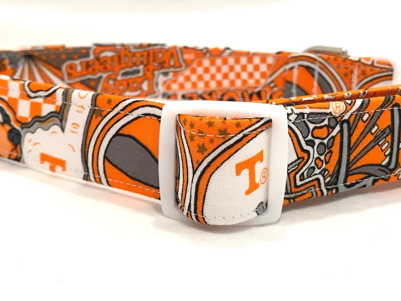 Puede incluir: Collar de perro naranja y gris con una hebilla blanca. El collar tiene un patr&oacute;n de estrellas, tableros de ajedrez y la letra "T" con un s&iacute;mbolo de marca registrada. El texto "Volunteer" tambi&eacute;n es visible en el collar.