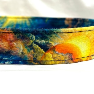 May include: A colorful dog collar with a blue, yellow, orange, and red abstract pattern. The collar is made of fabric and has a black stitched seam.