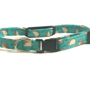 May include: A turquoise blue cat collar with a pattern of brown and white tacos. The collar has a black plastic buckle and a silver D-ring.