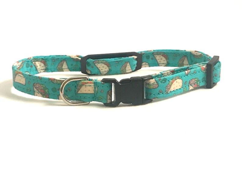 May include: A turquoise blue cat collar with a pattern of brown and white tacos. The collar has a black plastic buckle and a silver D-ring.