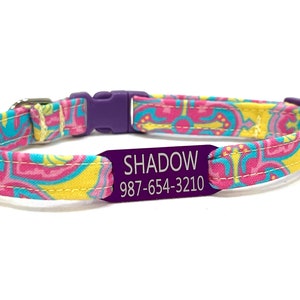 May include: Purple pet collar with a pink, yellow, and blue floral pattern. The collar has a purple engraved tag that reads "SHADOW 987-654-3210".