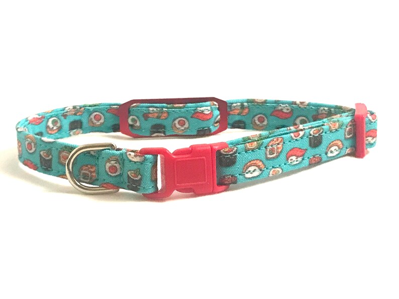 May include: A turquoise blue cat collar with a red buckle and a silver D-ring. The collar is decorated with a repeating pattern of sushi rolls.