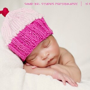 May include: A sleeping newborn baby wearing a knitted cupcake hat. The hat is pink and white with a red cherry. The baby is resting on a white blanket. The image has the text "Squid Inc. Studios Photography | Newborn".