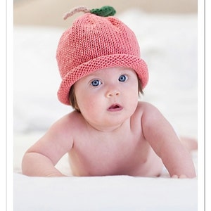 May include: A baby wearing a pink knitted hat shaped like a peach, with a green leaf and a tan stem. The baby has blue eyes and is lying on a white surface. The image is a portrait.