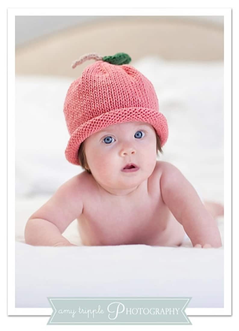 May include: A baby wearing a pink knitted hat shaped like a peach, with a green leaf and a tan stem. The baby has blue eyes and is lying on a white surface. The image is a portrait.