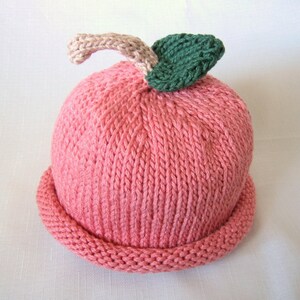 May include: A knitted baby hat resembling a peach. The hat is predominantly pink with a rolled brim. A beige stem and green leaf are attached to the top, completing the fruit design. The hat is made of soft yarn.