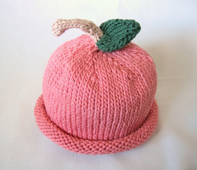 May include: A knitted baby hat resembling a peach. The hat is predominantly pink with a rolled brim. A beige stem and green leaf are attached to the top, completing the fruit design. The hat is made of soft yarn.