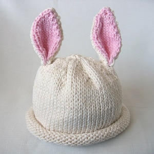 May include: A cream-coloured knitted hat with two upright bunny ears. The inside of the ears is pink. The hat has a rolled brim and is made of a soft, textured yarn. A cute accessory for babies and toddlers.
