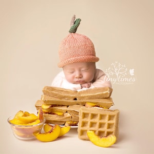 May include: A sleeping baby wearing a peach-themed knitted hat, resting on a stack of waffles. Sliced peaches are arranged in a glass bowl and on a plate. The image has a soft, warm color palette.