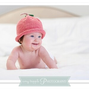 May include: A baby wearing a pink knitted hat with a green stem, smiling on a white surface. The hat resembles a strawberry. The image has soft lighting. The baby has fair skin and blue eyes. The background is light-coloured.