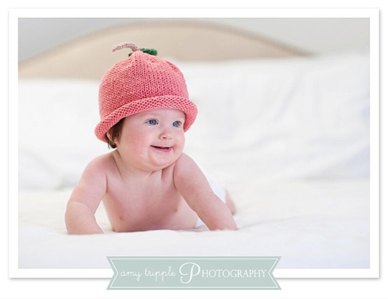 May include: A baby wearing a pink knitted hat with a green stem, smiling on a white surface. The hat resembles a strawberry. The image has soft lighting. The baby has fair skin and blue eyes. The background is light-coloured.