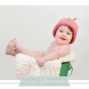 May include: A smiling baby wearing a pink knitted hat with a small stem detail, sitting in a green bucket lined with fluffy white material. The baby's legs are extended, and the background is a plain white.