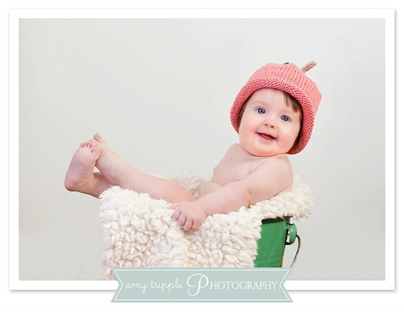 May include: A smiling baby wearing a pink knitted hat with a small stem detail, sitting in a green bucket lined with fluffy white material. The baby's legs are extended, and the background is a plain white.