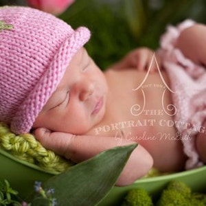 May include: A newborn baby wearing a pink and green knitted hat with a flower design, sleeping in a green basket. The baby is wearing a pink ruffled diaper cover.