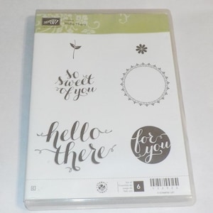 Può includere: Un set di sei timbri in gomma trasparente con diverse frasi e disegni, tra cui "so sweet of you", "hello there", "for you", un fiore, una foglia e un cerchio decorativo. I timbri sono confezionati in una custodia di plastica trasparente con il testo "Hostess Hello There" e "Stampin' Up!" stampato sul davanti.