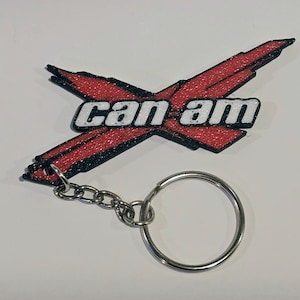 May include: A red and black glittery Can-Am keychain. The keychain features a stylized X design with the words "Can Am" in white. The keychain has a silver metal chain and key ring.