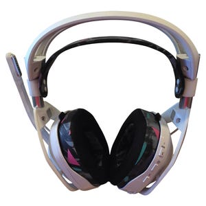 May include: White and black gaming headset with a microphone. The earcups have a black, pink, and teal geometric pattern. The headband is white with a black padded section. The microphone is on the left side.