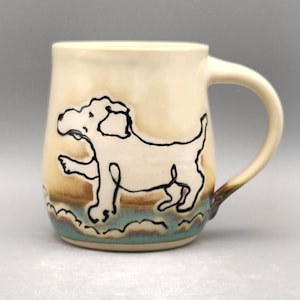 May include: Cream-colored ceramic mug with a black outline drawing of a dog. The mug has a curved handle and a base with blue and brown accents. The dog illustration is playful and whimsical.