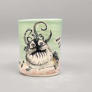 May include: A ceramic tumbler with a hand-drawn illustration of a whimsical sea creature. The creature is black and white with large eyes and a toothy grin, set against a light green background. The words "Deep Sea" are written on the side.