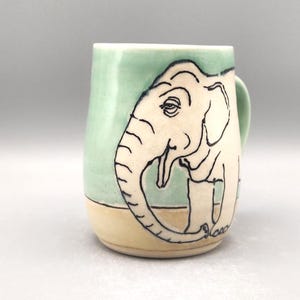 May include: Ceramic mug with a hand-drawn elephant design. The mug features a light green background with a cream-colored elephant outlined in black. The bottom of the mug is a light tan color. The mug has a handle and is suitable for hot or cold beverages.