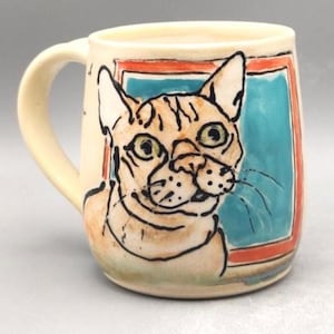 May include: Cream-colored ceramic mug featuring a hand-painted cat portrait. The cat has orange and black markings, green eyes, and is set against a blue background framed in red. The mug has a curved handle.