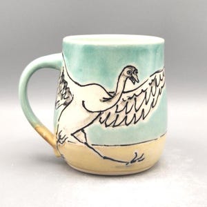 May include: Ceramic mug with a hand-painted design of a white bird in flight. The mug features a light blue background, a yellow base, and a light green handle. The bird is outlined in black, adding contrast to the design.