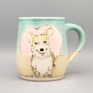 May include: A ceramic mug with a hand-painted corgi illustration. The mug features a light blue top, a beige bottom, and a pink heart-shaped background behind the corgi. The handle is light blue and beige.