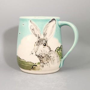 A close-up portrait of a hare with some leaping hares in the distance enhance this handmade, handpainted, unique ceramic mug