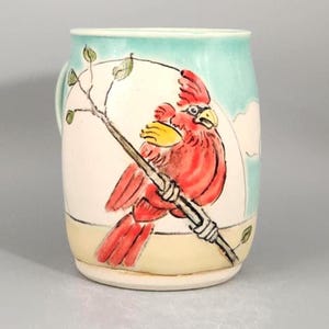 May include: Ceramic mug with a hand-painted cardinal bird design. The red bird is perched on a branch, set against a cream-colored background with a light blue sky. The mug has a rounded shape and a small handle.
