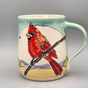 May include: Hand-painted ceramic mug featuring a vibrant red cardinal perched on a branch. The mug has a light blue handle and rim, with a white background behind the bird. The mug is approximately 4 inches tall.