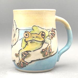 May include: A ceramic mug featuring a hand-painted illustration of a green frog with large eyes and orange-tipped toes. The mug has a cream-colored upper section, a light blue base, and a matching light blue handle, with a white background.