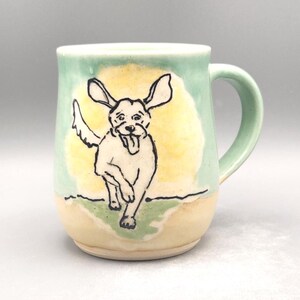 May include: Ceramic mug with a hand-drawn illustration of a running dog. The mug features a light green upper section, a yellow background behind the dog, and a cream-colored base. The dog is white with black outlines.